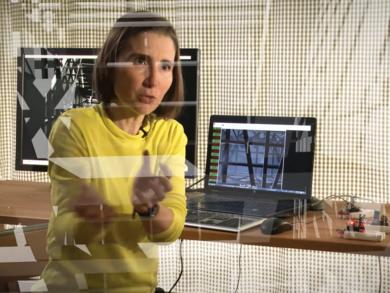 Frau in gelbem Pullover gestikuliert mit den Händen vor zwei Computerbildschirmen, an die kleine Roboter angeschlossen sind.