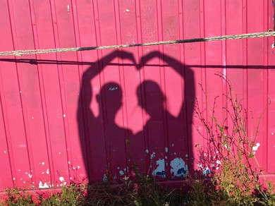 Schatten zweier Personen, die vor einer roten Wand mit ihren Armen ein Herz formen.