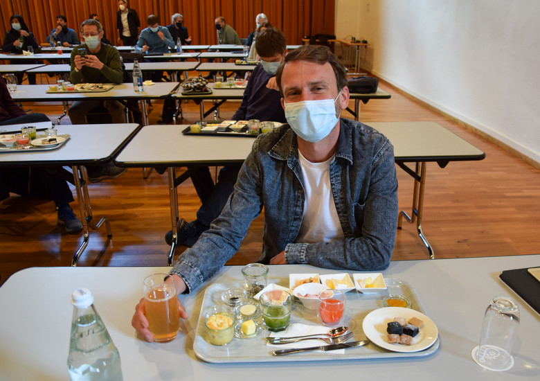 Junger Mann in Jeanshemd trägt eine Gesichtsmaske und sitzt in einer Mensa am Tisch; vor sich ein Tablett voller Essen und Getränke.