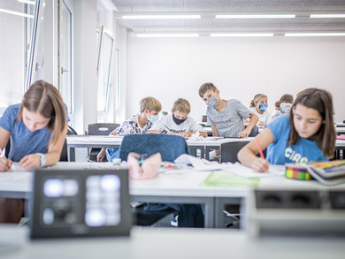 Schüler*innen bei der Arbeit im Klassenzimmer; manche tragen Gesichtsmasken.