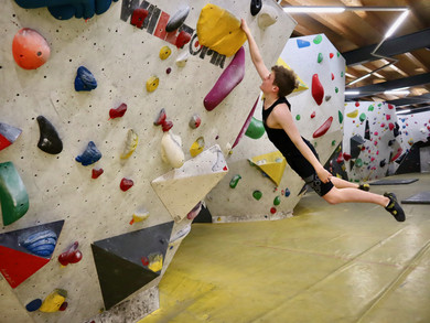 Blonder junger Mann an einer Boulderwand: Hält sich mit einer Hand an einem Griff fest, sein Körper ist weit von der Wand weg; er scheint zu fliegen.