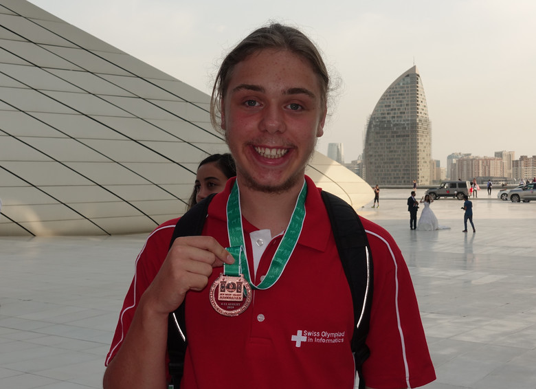 Junger, blondhaariger Mann mit Bart schaut lächelnd in die Kamera. Um seinen Hals hängt eine Medaille der Swiss Olympiad in Informatics.