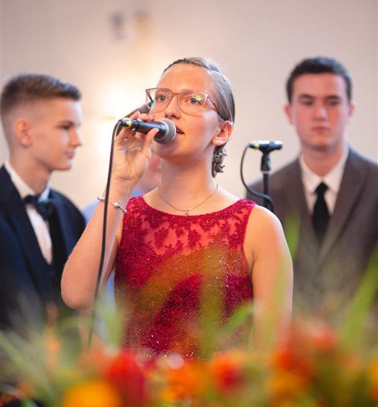 Blonde, junge Frau mit Brille und in rotem Kleid singt in Mikrofon.
