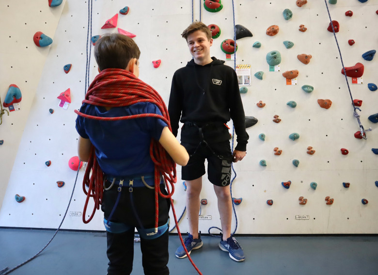 Blonder Junge vor Kletterwand, vor ihm ein kleinerer Junge von hinten mit Kletterseil um den Hals.