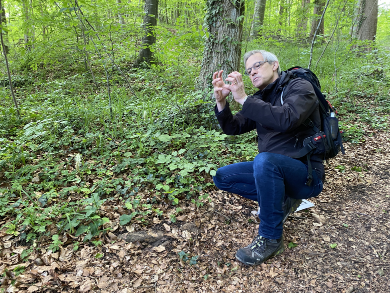Mann mit weissen Haaren und Brille kauert im Wald am Boden und formt die Hände vor seinem Gesicht zu einem Kreis, während er die Pflanzenwelt des Ittinger Waldes erklärt.