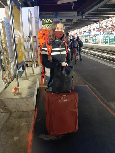 Eine junge Frau mit Maske und Koffern steht am Bahnhof.