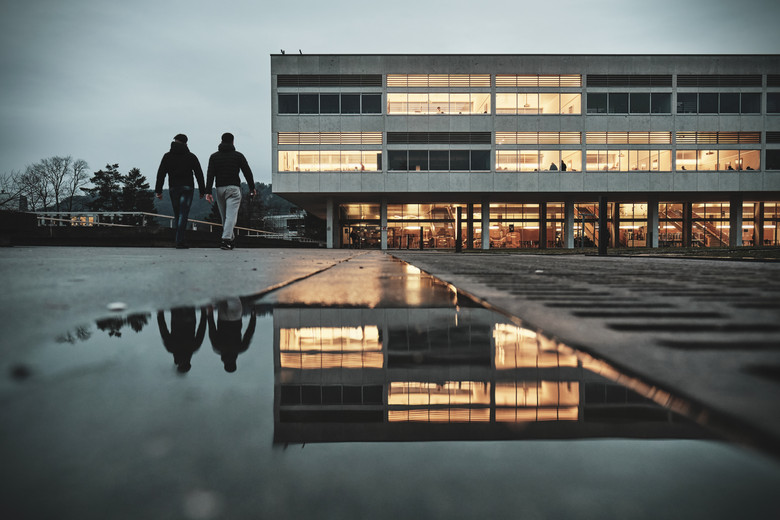 Ein Schulgebäude im brutalistischen Stil, davor zwei Schüler mit dem Rücken zu uns. Ein paar Fenster sind hell erleuchtet, das Licht spiegelt sich im Wasser am Boden.