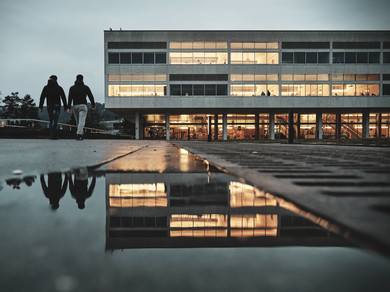 Ein Schulgebäude im brutalistischen Stil, davor zwei Schüler mit dem Rücken zu uns. Ein paar Fenster sind hell erleuchtet, das Licht spiegelt sich im Wasser am Boden.