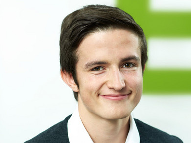 Junger dunkelhaariger Mann lächelt vor grün-weissem Hintergrund in die Kamera.
