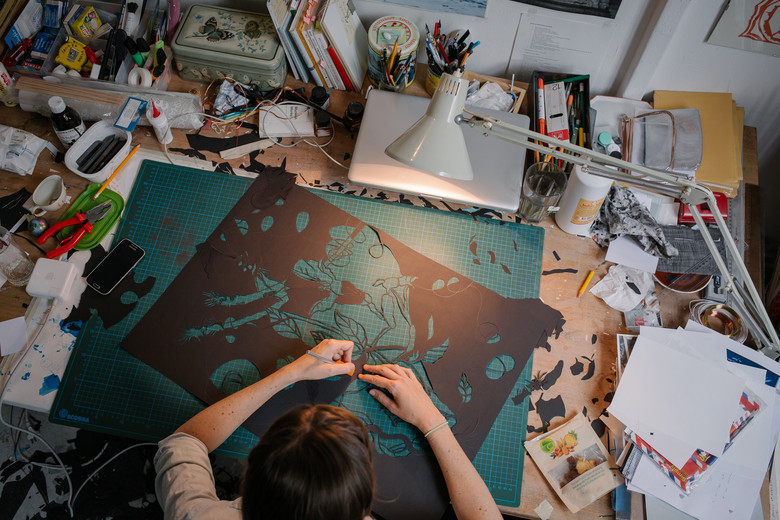 Künstlerin von oben fotografiert an einem Tisch mit Papier und Skalpell.