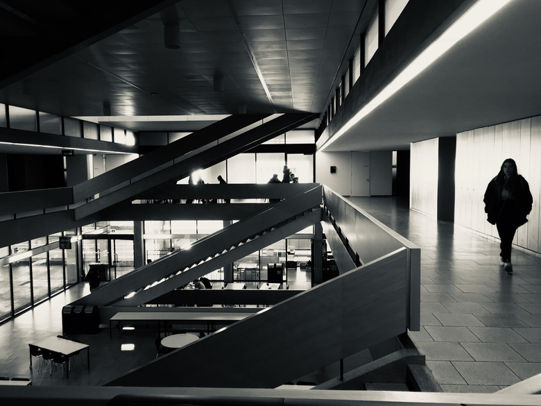 Ein Schwarzweiss-Bild eines Schulgebäudes im brutalistischen Stil, davor zwei Schüler mit dem Rücken zu uns. Ein paar Fenster sind hell erleuchtet, das Licht spiegelt sich im Wasser am Boden.