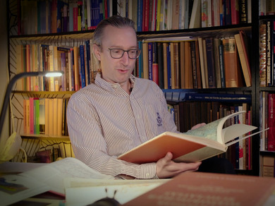 Marco Zanoli sitzt vor dem Bücherregal und liest im Atlas. 