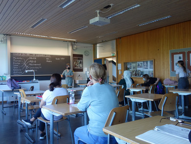 Ein Schulzimmer mit rund einem Dutzend Personen. An der Wandtafel stehen verschiedene Namen. 