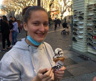 Eine junge Frau hält ein Glacé in der Hand, sie steht in der Fussgängerzone einer Stadt. 