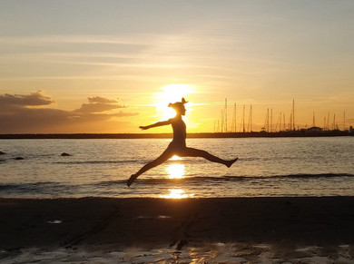 Eine Frau macht am Strand vor dem Sonnenuntergang einen Spagat in der Luft.