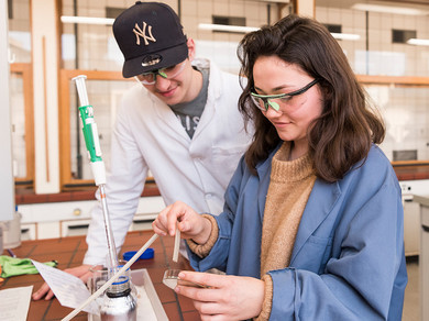 Drei Schüler*innen, in Labormäntel gekleidet, experimentieren im Labor während des NaTech-Unterrichtes.