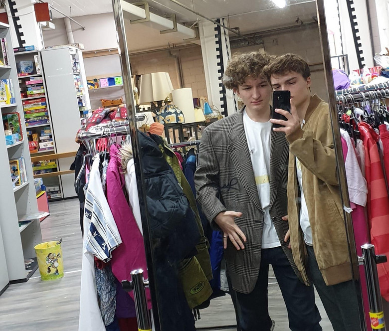 Zwei junge Männer probieren vor einem Spiegel Kleider an.