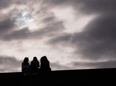 Vier Schüler*innen sitzen auf der Treppe eines Schulhauses vor einem bewölkten Himmel.