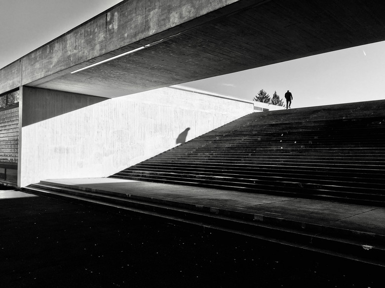 Ein Schwarzweiss-Foto einer grossen Betontreppe; zuoberst steht eine Person im Gegenlicht.