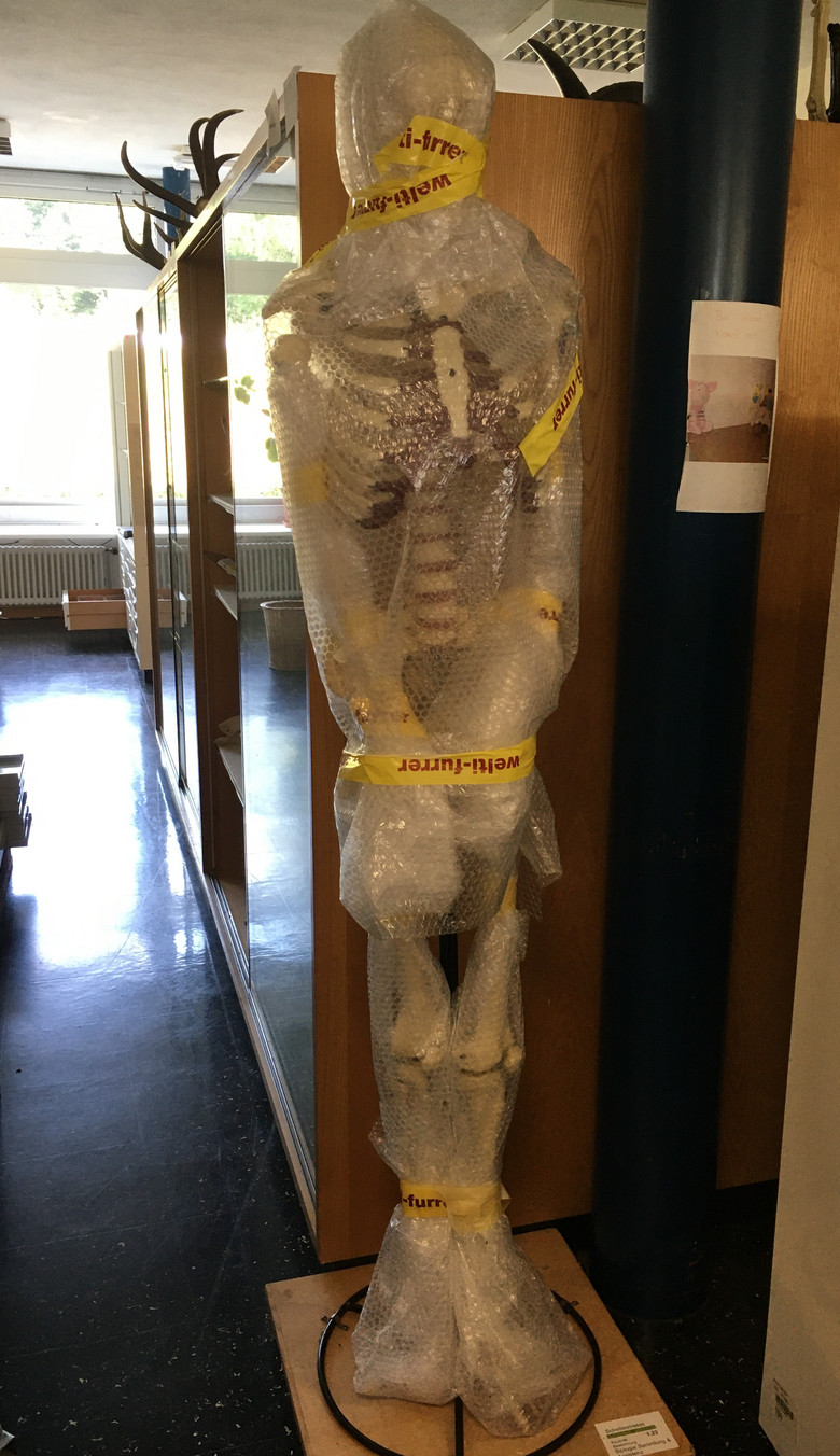 Ein mit Luftpolsterfolie umwickeltes Skelett steht in einem leeren Zimmer.
