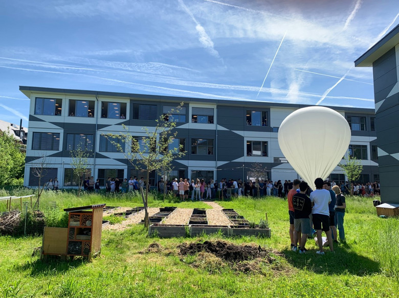Eine güne Wiese, darauf Schüler*innen mit einem grossen weissen Ballon und im Hintergrund ein Schulgebäude.