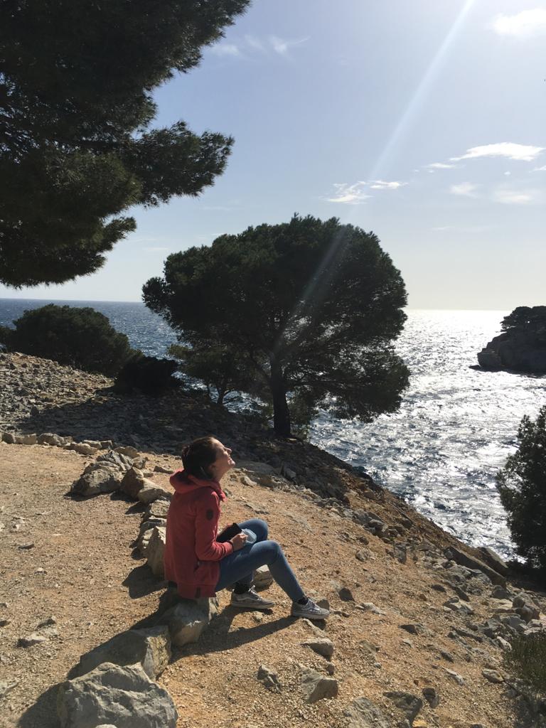 Eine junge Frau sitzt auf den Felsen am Meer und schaut in die Sonne.